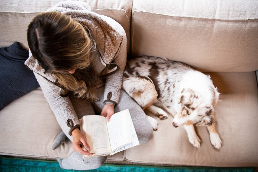 woman in K&Uuml;HL Flight Jacket reading book on the couch next to white dog